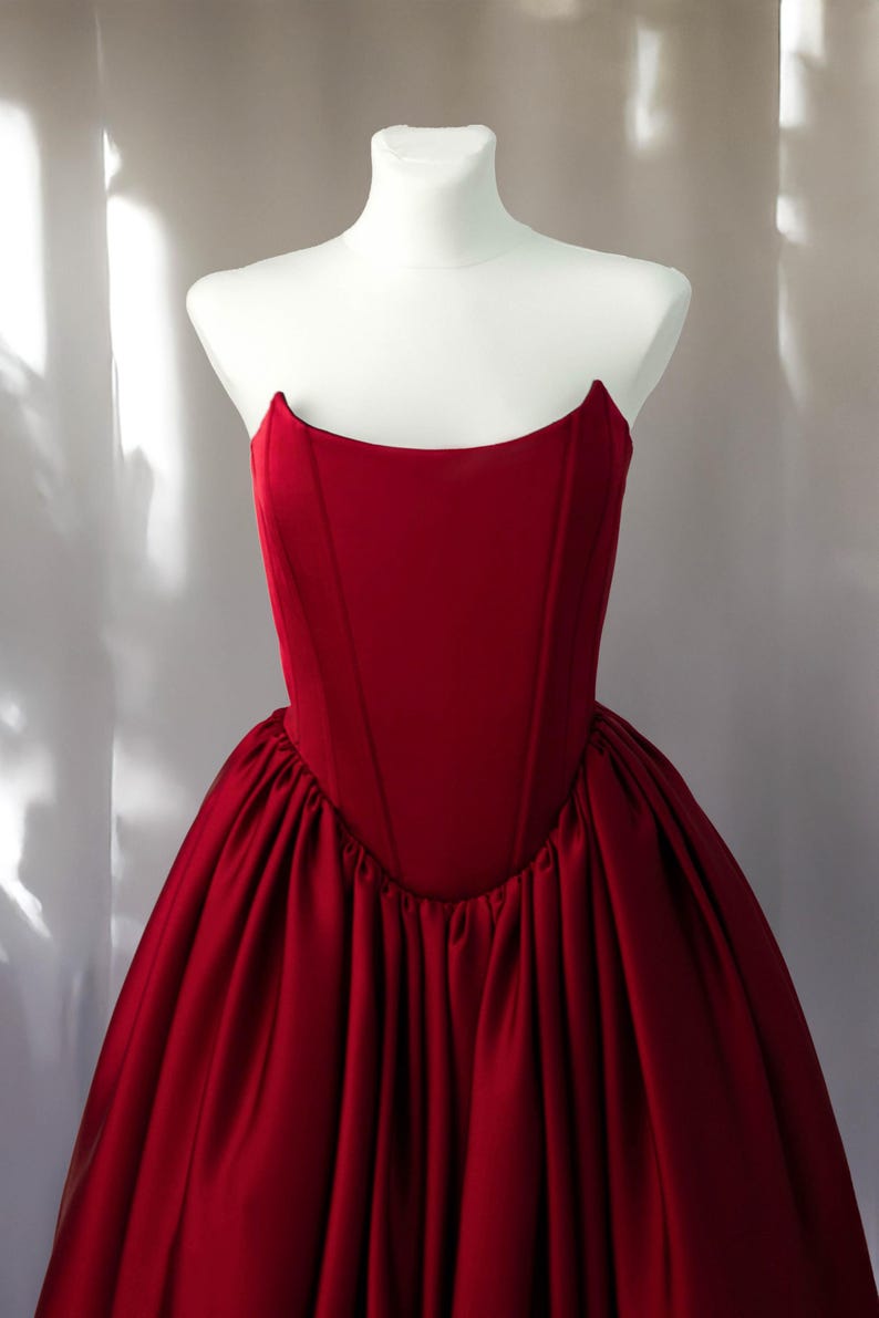 May include: A deep red strapless gown featuring a structured corset-style bodice with vertical seams and a voluminous, gathered skirt. The garment is displayed on a white mannequin, showcasing a smooth, matte finish on the bodice and fabric.