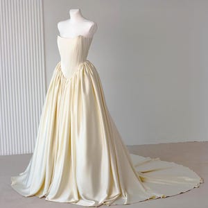 May include: A floor-length cream-coloured gown featuring a structured, strapless corset bodice with vertical boning details and a voluminous, gathered skirt that extends into a long train, displayed on a white mannequin against a neutral background.
