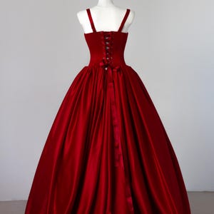 May include: A floor-length, deep red gown featuring a structured bodice with a lace-up corset back, thick shoulder straps, and a voluminous, full-length skirt made of a smooth, satin-like fabric that drapes down to the floor in soft folds.