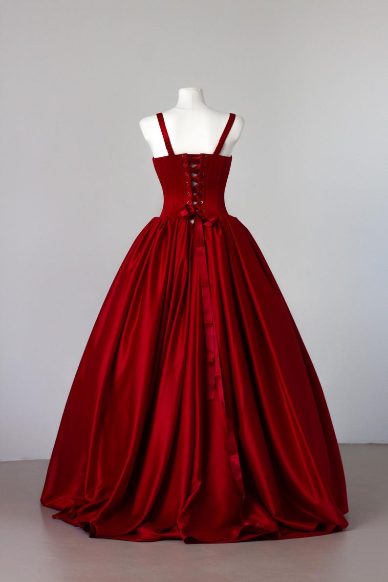 May include: A floor-length, deep red gown featuring a structured bodice with a lace-up corset back, thick shoulder straps, and a voluminous, full-length skirt made of a smooth, satin-like fabric that drapes down to the floor in soft folds.