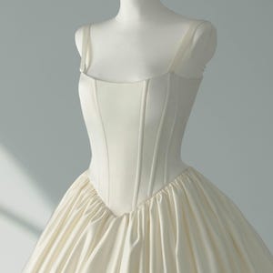 May include: An ivory-colored gown featuring a structured, corset-style bodice with vertical seams and a square neckline. The bodice transitions into a voluminous, gathered skirt made of smooth, satin-like fabric with a subtle sheen and soft folds.