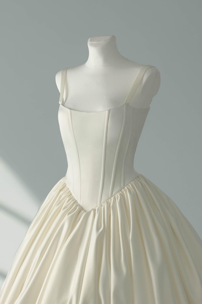 May include: An ivory-colored gown featuring a structured, corset-style bodice with vertical seams and a square neckline. The bodice transitions into a voluminous, gathered skirt made of smooth, satin-like fabric with a subtle sheen and soft folds.