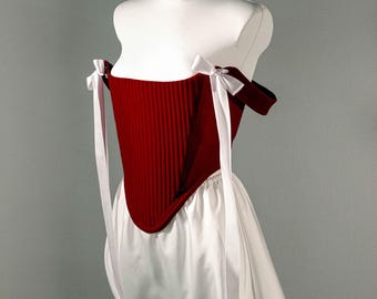 May include: A deep red corset featuring vertical stitched channels and off-the-shoulder sleeves with white ribbon ties at the shoulders. The garment is paired with a white skirt that has a gathered waistline, displayed on a white mannequin torso.