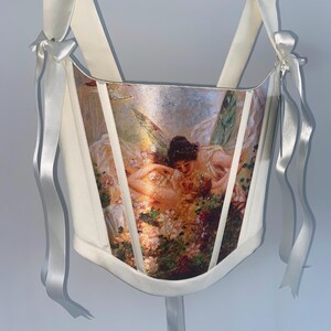 Puede incluir: Cors&eacute; color crema con estampado de pintura renacentista. El cors&eacute; presenta cintas y lazos de raso blanco. La obra de arte representa figuras en un jard&iacute;n con elementos florales.