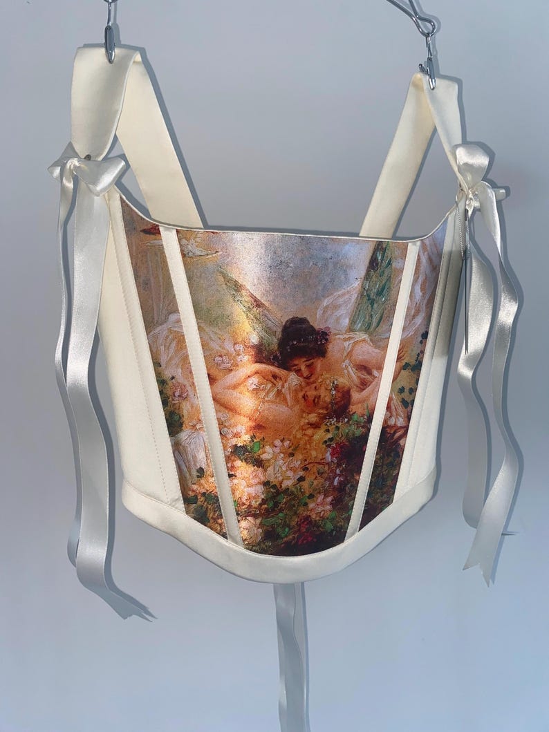 Puede incluir: Cors&eacute; color crema con estampado de pintura renacentista. El cors&eacute; presenta cintas y lazos de raso blanco. La obra de arte representa figuras en un jard&iacute;n con elementos florales.