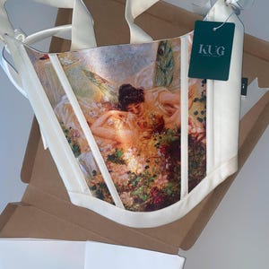 Puede incluir: Cors&eacute; color marfil con estampado de pintura rom&aacute;ntica, con flores y figuras. El cors&eacute; tiene correas y ribetes color marfil, y una etiqueta con "KUG". Presentado en una caja de cart&oacute;n.