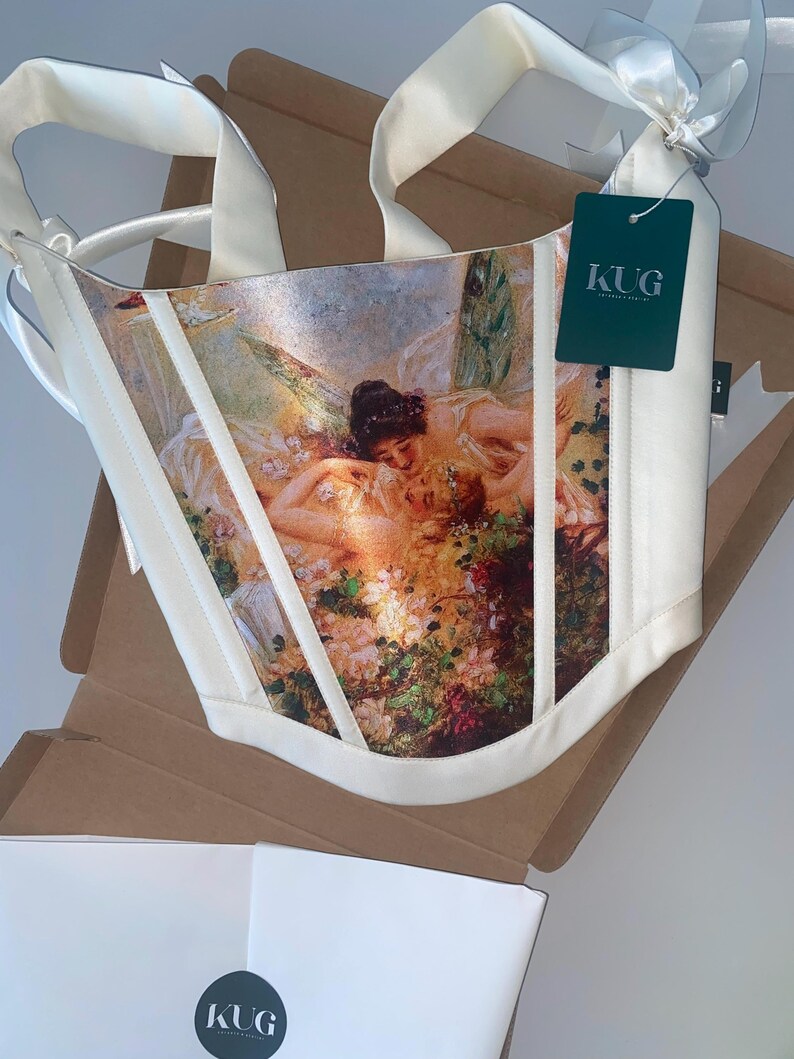 Puede incluir: Cors&eacute; color marfil con estampado de pintura rom&aacute;ntica, con flores y figuras. El cors&eacute; tiene correas y ribetes color marfil, y una etiqueta con "KUG". Presentado en una caja de cart&oacute;n.