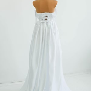 May include: A white, strapless gown with a corset back and flowing skirt. The dress is displayed on a mannequin with a tan-colored neck and shoulders. The gown's fabric drapes elegantly, creating a classic silhouette.