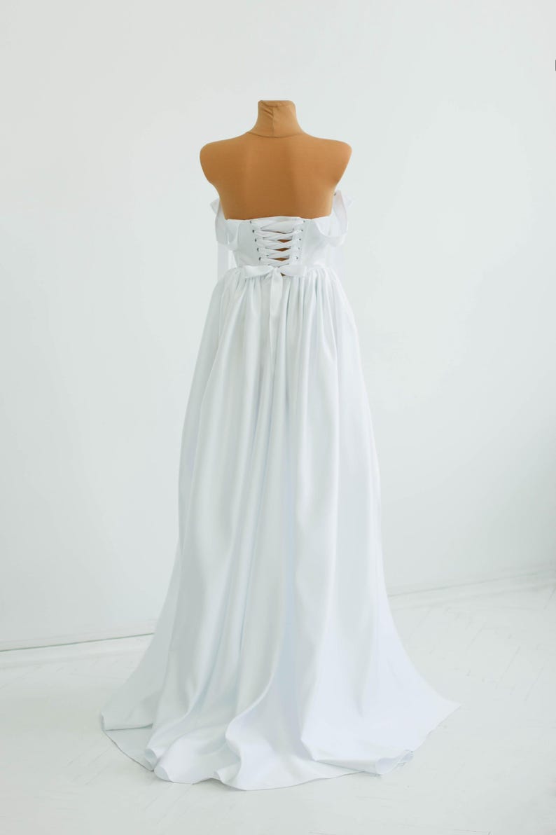 May include: A white, strapless gown with a corset back and flowing skirt. The dress is displayed on a mannequin with a tan-colored neck and shoulders. The gown's fabric drapes elegantly, creating a classic silhouette.