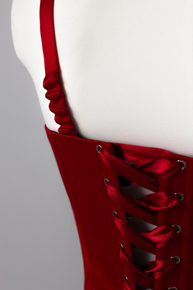 May include: A deep red corset top featuring a back lace-up closure with matching satin ribbon threaded through silver-toned metal grommets. The garment includes a wide shoulder strap with a gathered, ruched fabric texture along the top edge.