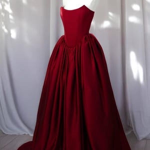 May include: A floor-length, deep red gown featuring a structured, strapless bodice with a sweetheart neckline and a voluminous, pleated skirt that cascades to the floor. The garment is made of a smooth, solid-colored fabric with a subtle sheen.