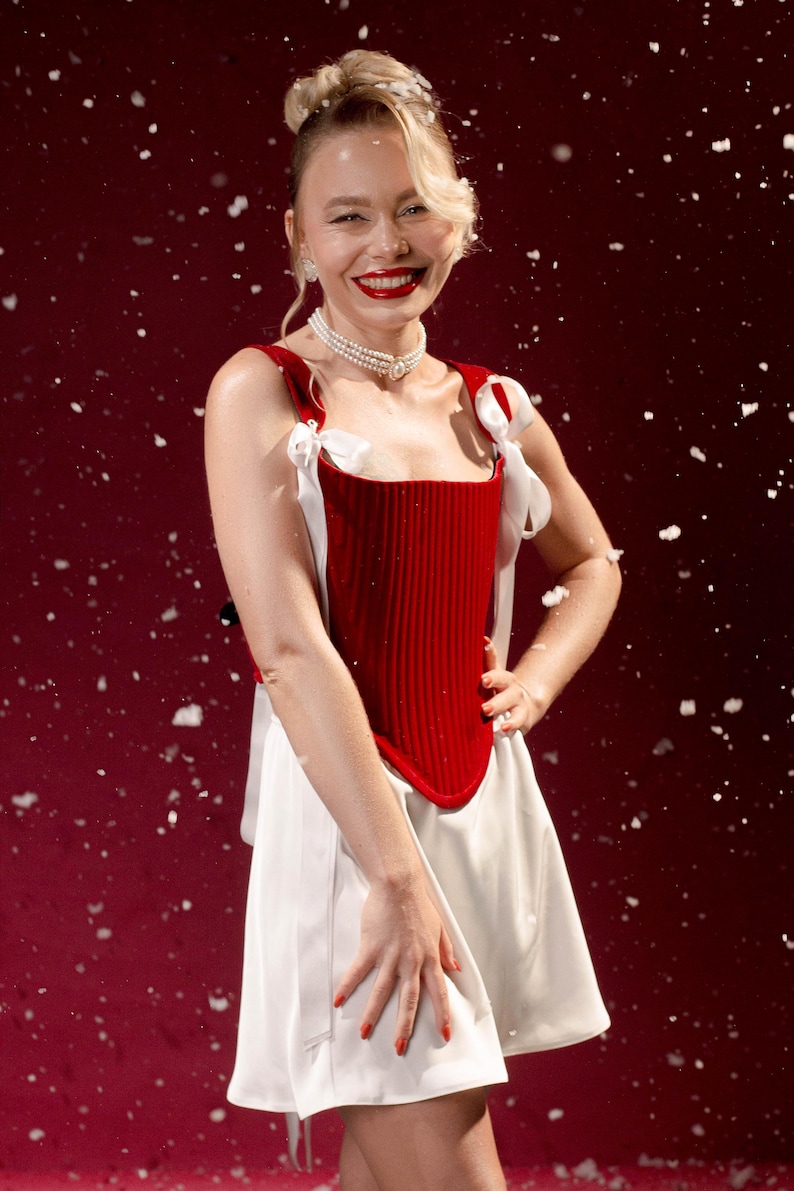 May include: A sleeveless dress featuring a deep red, vertically ribbed corset-style bodice with white ribbon shoulder ties and a flared white skirt. The garment is accessorized with a multi-strand pearl choker necklace against a dark red background.