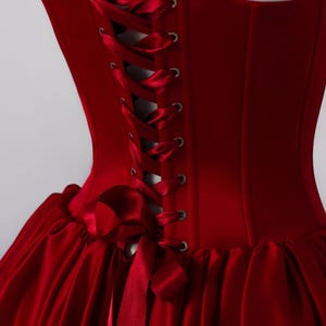 May include: A deep red corset dress featuring a structured bodice with silver-toned metal grommets and a matching satin ribbon lace-up back closure. The garment includes gathered shoulder straps and a full, pleated skirt made of smooth satin fabric.