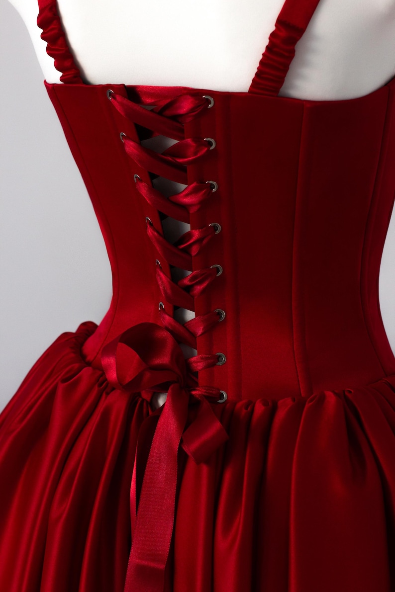 May include: A deep red corset dress featuring a structured bodice with silver-toned metal grommets and a matching satin ribbon lace-up back closure. The garment includes gathered shoulder straps and a full, pleated skirt made of smooth satin fabric.