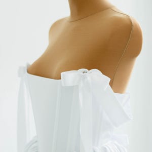 May include: A white, off-the-shoulder dress on a mannequin. The dress features a fitted bodice and a flowing skirt. White bows adorn the shoulders. The dress is made of a soft, flowing fabric, perfect for a special occasion.