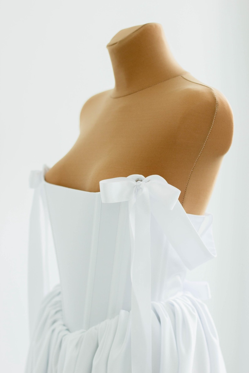 May include: A white, off-the-shoulder dress on a mannequin. The dress features a fitted bodice and a flowing skirt. White bows adorn the shoulders. The dress is made of a soft, flowing fabric, perfect for a special occasion.