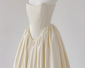 May include: A strapless cream-colored gown featuring a structured, corset-style bodice with vertical paneling and a voluminous, floor-length skirt with deep, gathered pleats that create a full, flowing silhouette from the waist down.