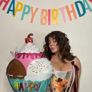 Puede incluir: Una colorida pancarta de "Happy Birthday" cuelga sobre una persona. Un gran globo con forma de helado con las palabras "Happy Birthday" se sostiene frente a la persona. El globo presenta secciones rosas, marrones y blancas con chispas.