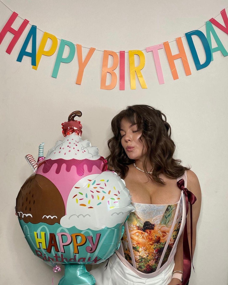 Puede incluir: Una colorida pancarta de "Happy Birthday" cuelga sobre una persona. Un gran globo con forma de helado con las palabras "Happy Birthday" se sostiene frente a la persona. El globo presenta secciones rosas, marrones y blancas con chispas.
