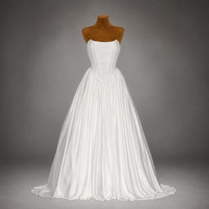 May include: A floor-length white strapless gown featuring a structured, corset-style bodice with vertical seams and a full, pleated skirt that flares out from the waist. The garment is displayed on a brown mannequin torso against a neutral background.