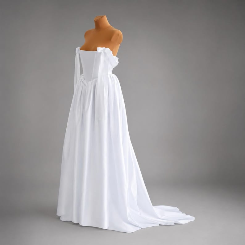May include: A white, off-the-shoulder gown with a flowing skirt and a slight train. The dress is displayed on a mannequin, showcasing its elegant design. The bodice features a structured, strapless design, and the skirt drapes gracefully to the floor.