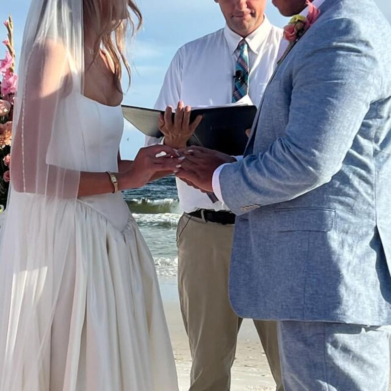 May include: A white wedding dress with a square neckline and a sheer veil, a light blue textured blazer, and a white button-down shirt with a blue plaid tie. The individuals are exchanging rings during an outdoor ceremony near the ocean.
