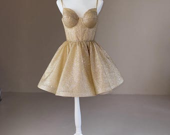 May include: A gold glittery mini dress featuring a structured corset-style bodice with molded cups, thin spaghetti straps, and a flared, pleated skirt. The garment is displayed on a white mannequin, showcasing a shimmering texture throughout.
