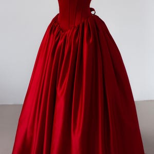 May include: A floor-length, deep red satin gown featuring a structured, sleeveless bodice with a square neckline and thick straps. The skirt is voluminous and gathered at the waist, creating a full, pleated silhouette that extends to the floor.