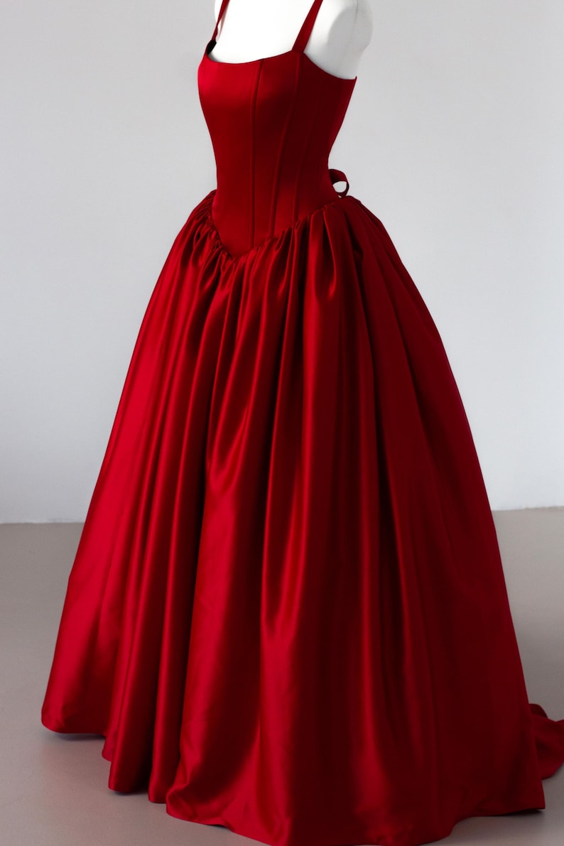 May include: A floor-length, deep red satin gown featuring a structured, sleeveless bodice with a square neckline and thick straps. The skirt is voluminous and gathered at the waist, creating a full, pleated silhouette that extends to the floor.