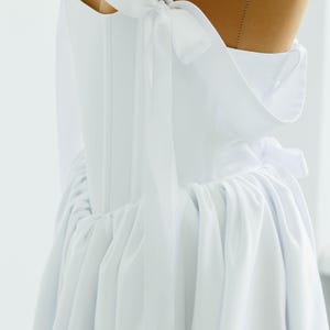 May include: A white dress with a corset-style bodice and a flowing skirt. The dress features delicate bow details on the shoulders and a gathered waistline. The fabric appears to be a lightweight material, creating a soft, elegant silhouette.