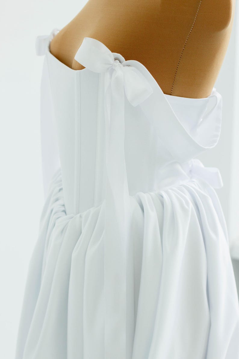 May include: A white dress with a corset-style bodice and a flowing skirt. The dress features delicate bow details on the shoulders and a gathered waistline. The fabric appears to be a lightweight material, creating a soft, elegant silhouette.