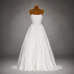 May include: A strapless white gown featuring a structured, corset-style bodice with vertical seams and a full, floor-length skirt with gathered fabric. The garment is displayed on a brown mannequin torso against a neutral grey background.