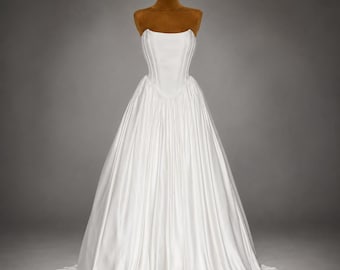 Puede incluir: Un vestido blanco sin tirantes que presenta un corpiño estructurado estilo corsé con costuras verticales y una falda larga y completa con tela fruncida. La prenda se exhibe en un torso de maniquí marrón sobre un fondo gris neutro.