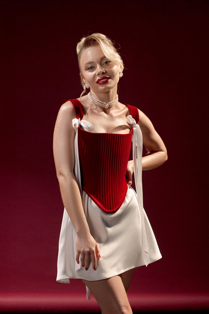 May include: A sleeveless dress featuring a deep red, vertically ribbed corset-style bodice with a pointed hemline. The bodice is accented with white ribbon shoulder ties. The skirt is a solid white, smooth fabric that flares slightly from the waist.