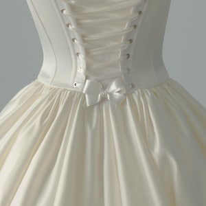 May include: An ivory-colored satin dress featuring a structured bodice with a lace-up back closure. The corset-style back includes metallic eyelets and a matching ribbon tie, finished with a small decorative satin bow at the waistline.