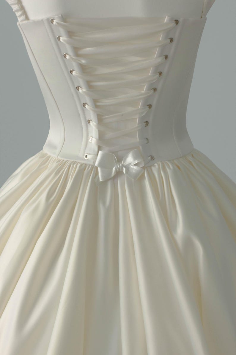 May include: An ivory-colored satin dress featuring a structured bodice with a lace-up back closure. The corset-style back includes metallic eyelets and a matching ribbon tie, finished with a small decorative satin bow at the waistline.