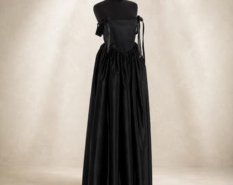 May include: A floor-length black dress featuring a fitted bodice with a straight neckline and thin ribbon shoulder ties. The skirt is voluminous and gathered at the waist, creating a full silhouette. The fabric has a subtle sheen throughout.