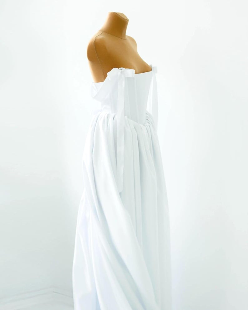 May include: A white off-the-shoulder gown with a flowing skirt. The bodice is fitted, and the dress features delicate tie details at the shoulders. The dress is displayed on a mannequin against a white background.