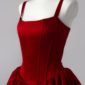 May include: A deep red satin dress featuring a structured bodice with vertical seams and wide shoulder straps. The garment has a fitted torso that transitions into a voluminous, gathered skirt with a smooth, reflective finish throughout the fabric.