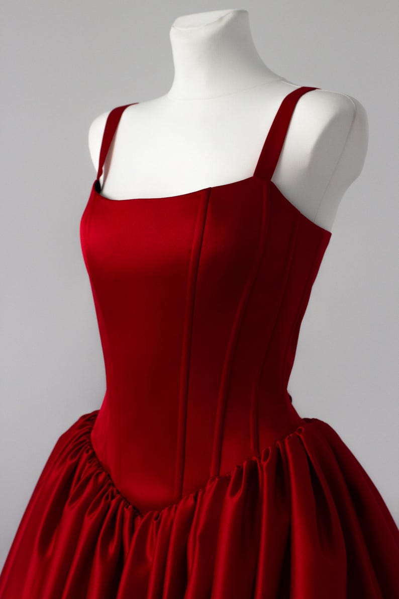 May include: A deep red satin dress featuring a structured bodice with vertical seams and wide shoulder straps. The garment has a fitted torso that transitions into a voluminous, gathered skirt with a smooth, reflective finish throughout the fabric.