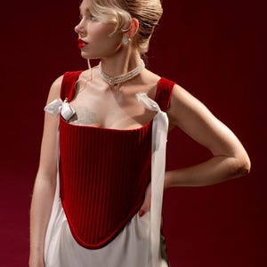 May include: A deep red velvet corset top featuring vertical ribbed texture, a pointed hemline, and white satin ribbon shoulder ties. The garment is paired with a white skirt and a multi-strand pearl choker necklace with a central circular pendant.