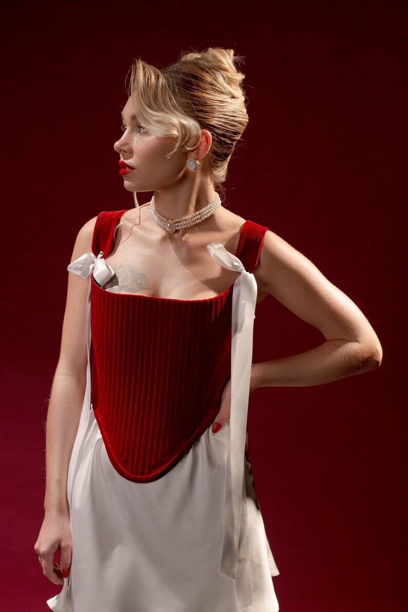 May include: A deep red velvet corset top featuring vertical ribbed texture, a pointed hemline, and white satin ribbon shoulder ties. The garment is paired with a white skirt and a multi-strand pearl choker necklace with a central circular pendant.