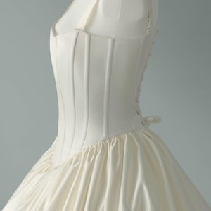 May include: An ivory-colored gown featuring a structured, boned bodice with vertical seams and a square neckline. The bodice connects to a voluminous, gathered skirt. The side of the garment displays a row of small, matching fabric-covered buttons.