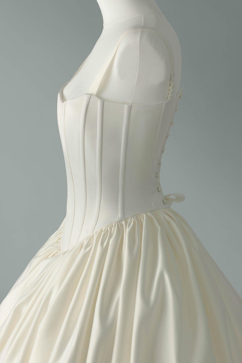 May include: An ivory-colored gown featuring a structured, boned bodice with vertical seams and a square neckline. The bodice connects to a voluminous, gathered skirt. The side of the garment displays a row of small, matching fabric-covered buttons.