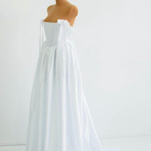 May include: A white, strapless wedding dress on a mannequin. The dress features a fitted bodice and a flowing skirt that extends into a train. Delicate white ribbons are tied at the shoulders. The dress is displayed against a plain white background.