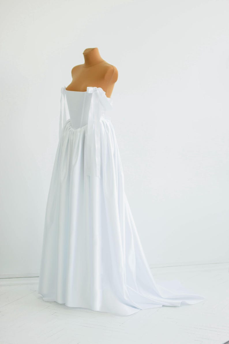 May include: A white, strapless wedding dress on a mannequin. The dress features a fitted bodice and a flowing skirt that extends into a train. Delicate white ribbons are tied at the shoulders. The dress is displayed against a plain white background.