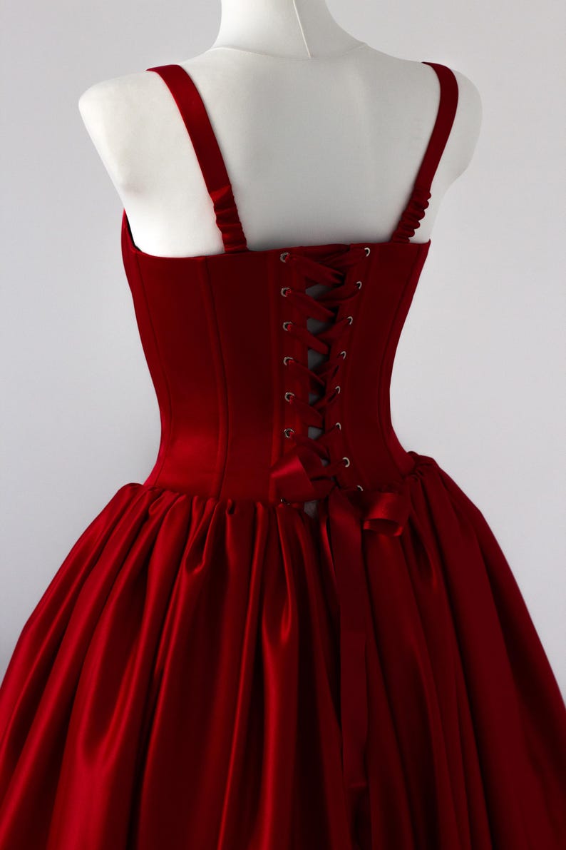 May include: A deep red corset dress featuring a structured bodice with a lace-up back closure using matching ribbon, wide shoulder straps, and a full, gathered skirt. The garment is made of a smooth, solid-colored fabric with a subtle sheen.