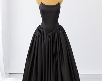 May include: A floor-length black gown featuring a structured, strapless corset-style bodice with vertical seams and a voluminous, gathered skirt that extends into a train. The garment is made of a smooth, solid black fabric with a subtle sheen.