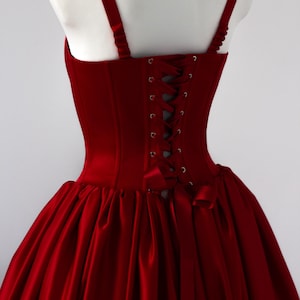 May include: A deep red corset dress featuring a structured bodice with a lace-up back closure using matching ribbon, wide shoulder straps, and a full, gathered skirt. The garment is made of a smooth, solid-colored fabric with a subtle sheen.