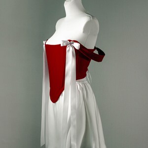 May include: A red corset-style top with white ribbon ties and a white skirt. The top has a structured design with a sweetheart neckline and is paired with a flowing skirt. The outfit is displayed on a mannequin.
